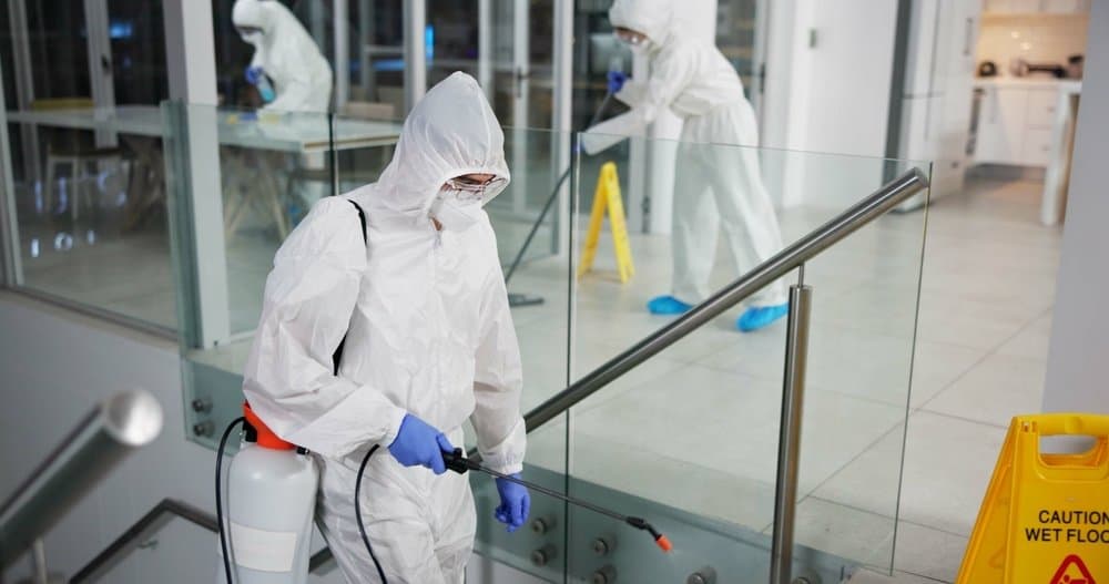 Workers in white protective suits treating an office interior, one in the foreground spraying beside a glass stair balustrade