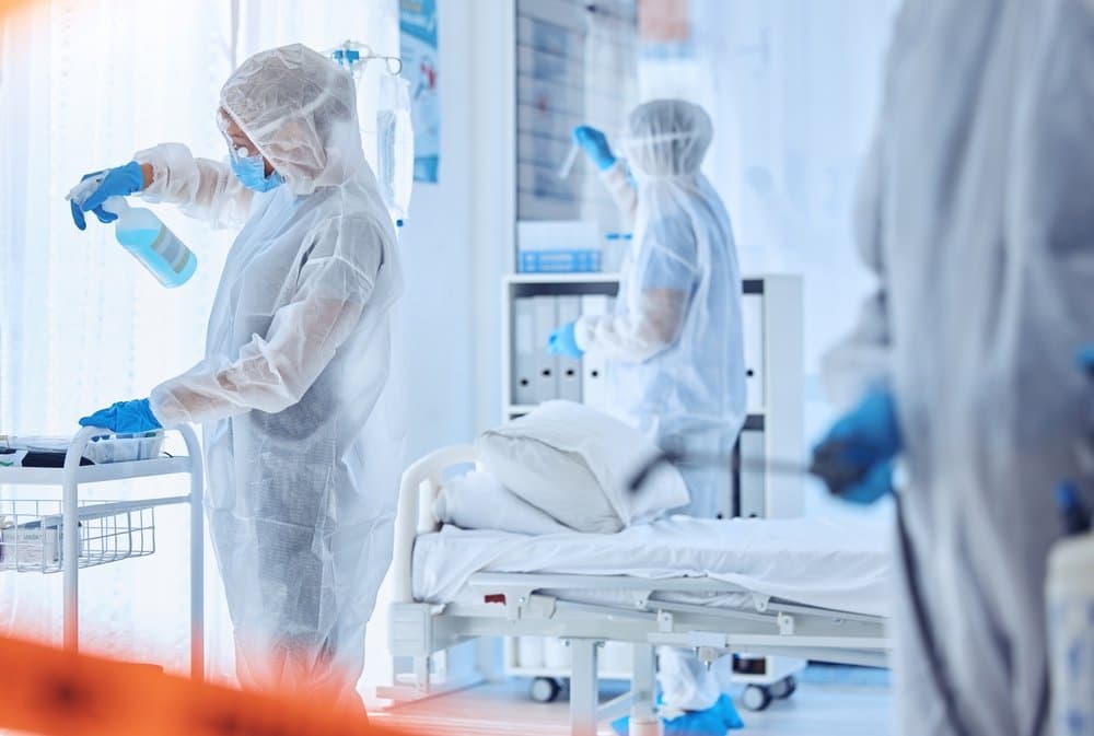 Three workers in white protective suits, masks and gloves disinfecting a hospital room, one spraying a trolley beside a bed