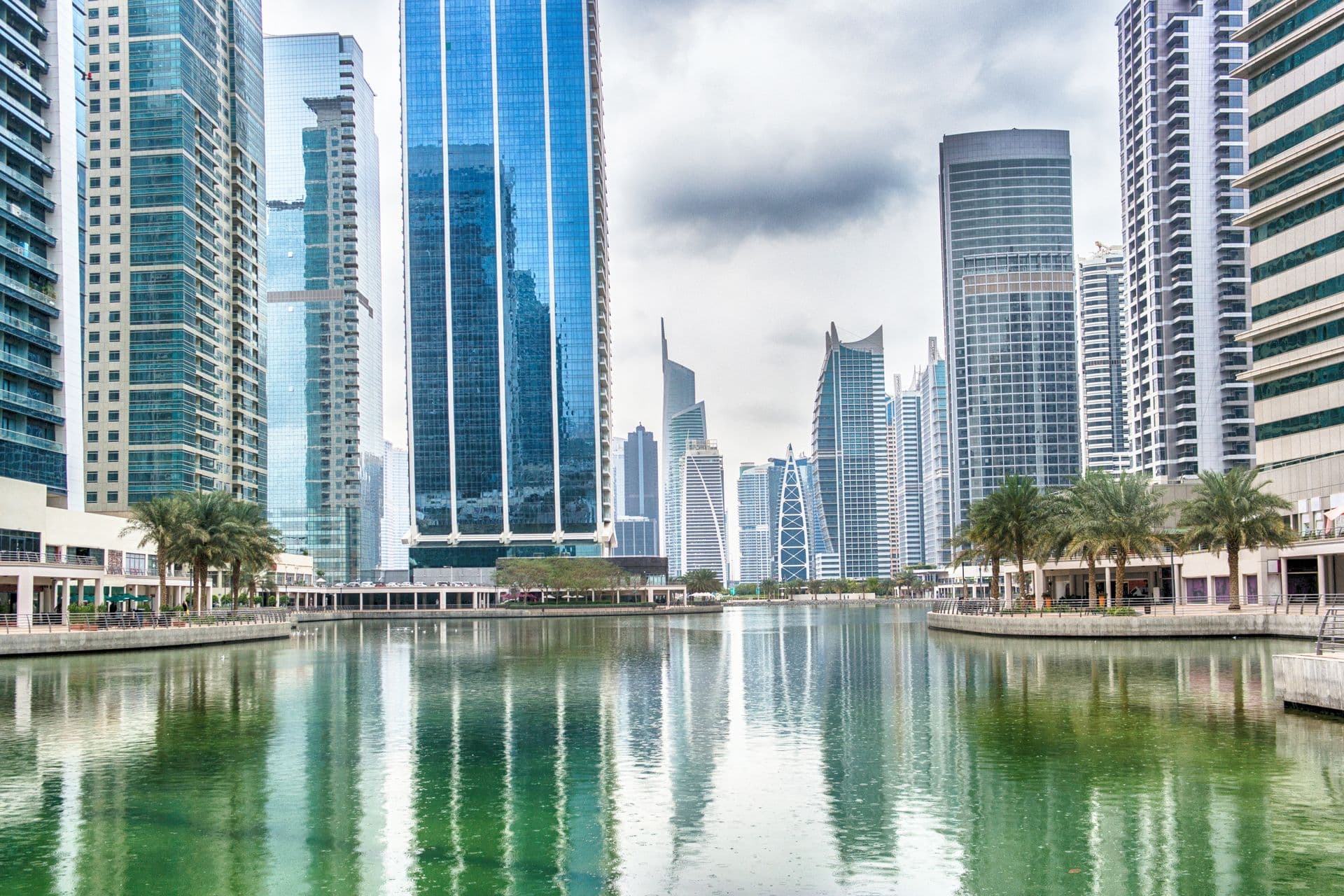 Glass towers in Jumeirah Lake Towers reflected in one of the lakes, with palm trees along the promenade