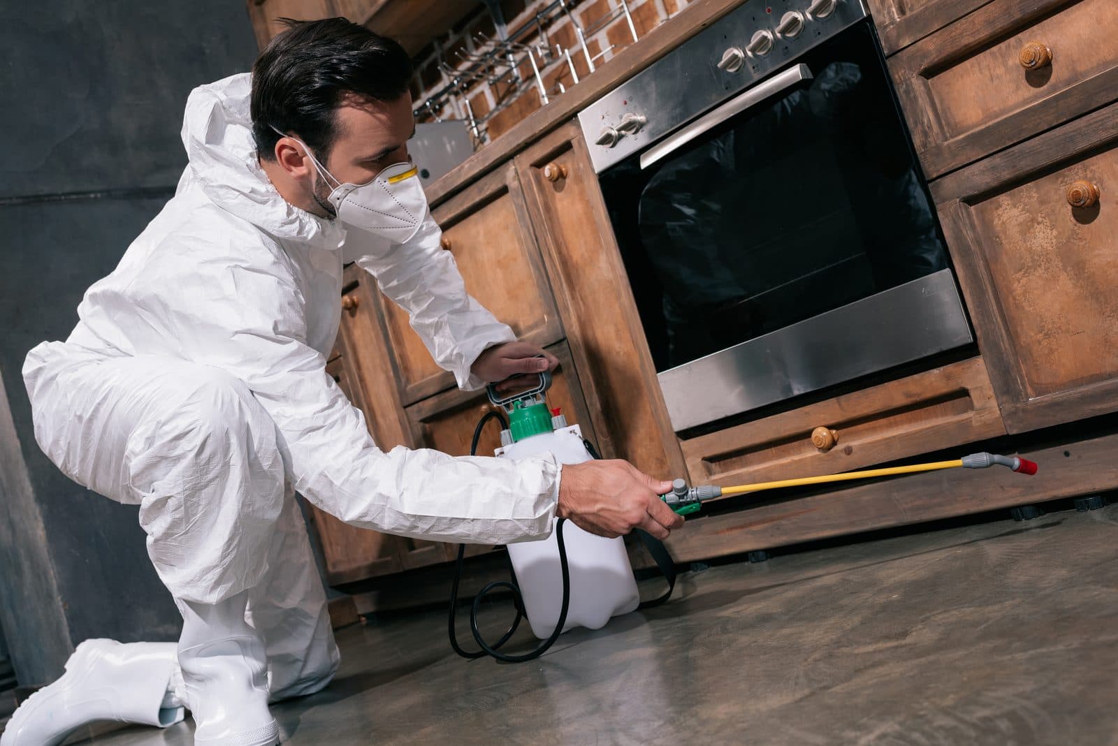 Technician in a white protective suit and face mask crouching to spray along the base of kitchen cabinets beside an oven