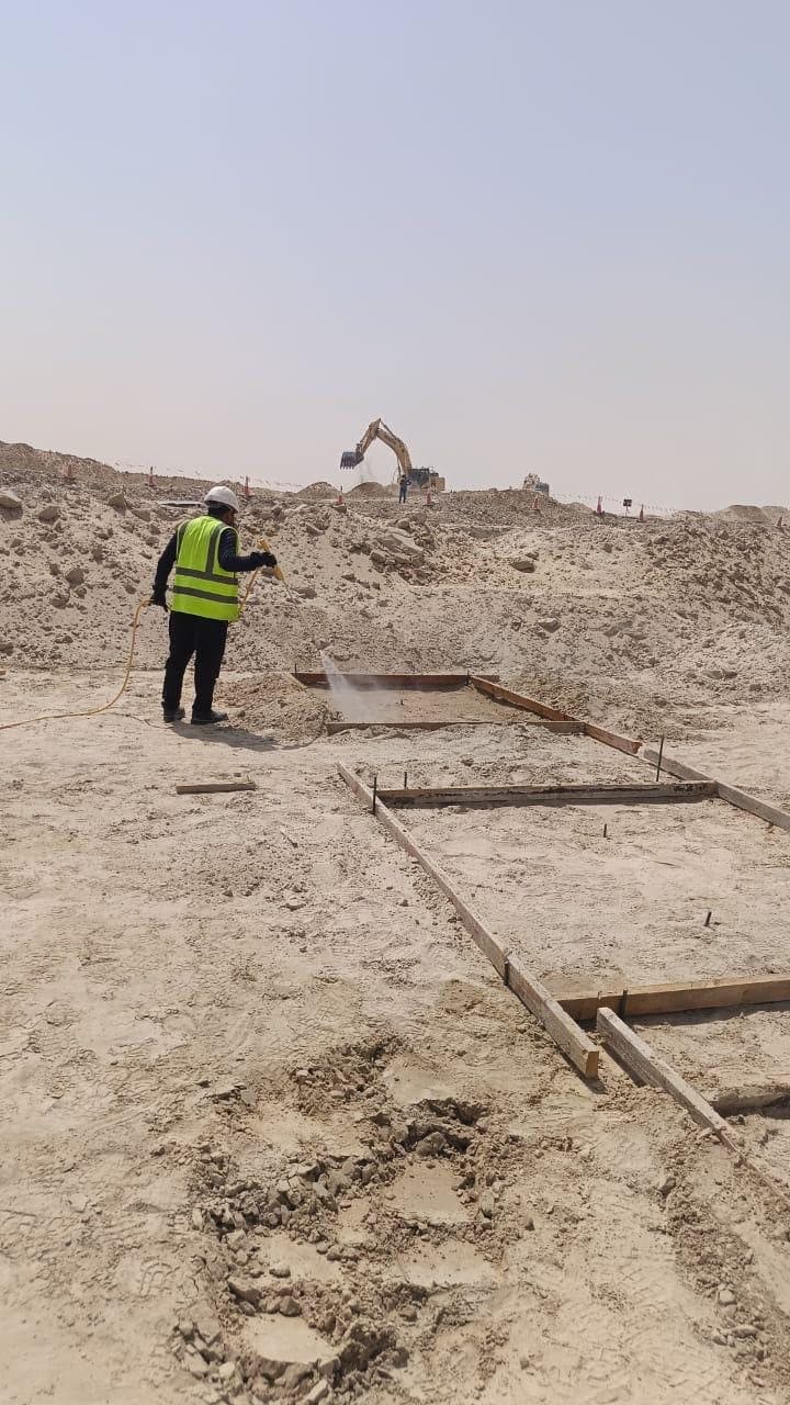 Technician treating prepared ground at the edge of a foundation excavation, an excavator working on the spoil heap beyond