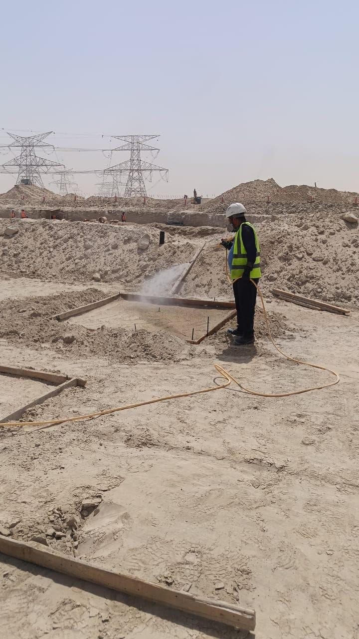 Technician in a hi-vis vest, hard hat and respirator spraying termiticide into a timber-formed footing excavation, power pylons on the horizon behind