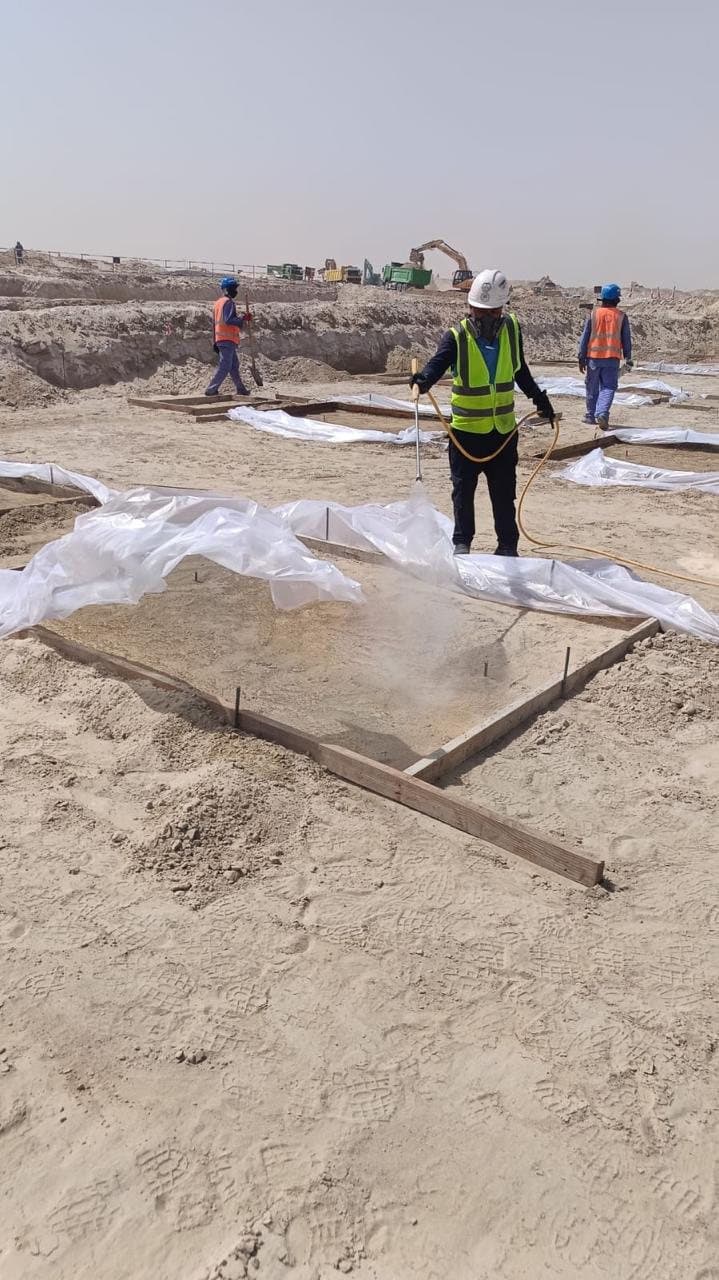 Technician applying an anti-termite soil barrier beside polythene-covered footings while two colleagues in hi-vis work further up the excavation